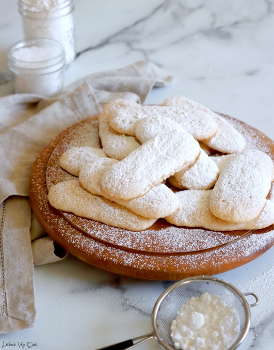 Pile of ladyfinger cookies, topped with sprinkled icing sugar, on a round wood board with white-grey marble background. A small metal sifter with icing sugar sits in front of the board and there's a light brown towel to the left with two small jars of white ingredients blurred in the back left corner.