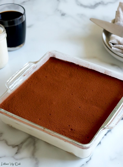 Top view of a square glass baking dish filled with tiramisu, with a layer of cream visible under a layer of cocoa powder. Tiramisu on a white-grey marble background with a glass mug of black coffee and small jar of milk to the top left corner and stack of plate with a folded towel at top right corner.