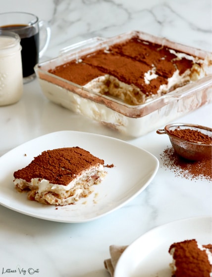 White plate with square of vegan tiramisu (layers of vanilla ladyfingers, cream and cocoa powder on top). A second plate with tiramisu slice in bottom right corner with a small metal sifter of cocoa powder to the center right of image. Square glass dish of sliced tiramisu in back with coffee and milk in glass mugs.