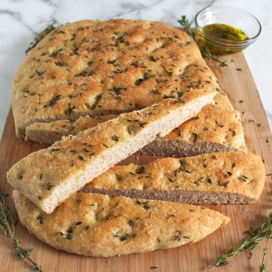 Large loaf of sliced focaccia bread with a wedge of bread resting on top, sitting on wooden cutting board with small jar of olive oil to back right and fresh thyme springs place around bread