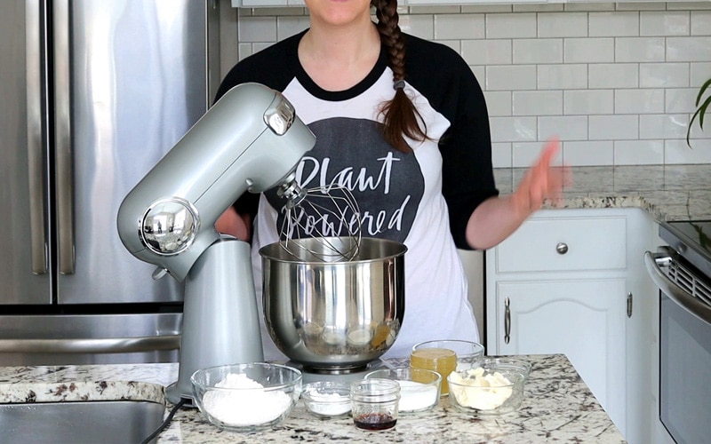 Grey stand mixer with whisk attachment on granite countertop surrounded by small glass bowls of ingredients (flour, corn starch, vanilla extract, butter, sugar and aquafaba are visible). Nicole stands behind the counter but her head is cropped out.