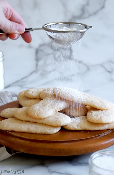 Hand holding a small metal sifter filled with icing sugar, sprinkling sugar onto a pile of ladyfinger biscuits sitting on a round wood board, with white-grey marble background.