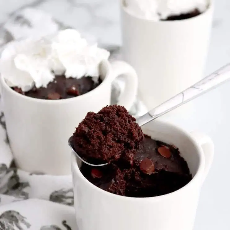 Three white mugs filled with microwaved brownie; two mugs in back topped with whip cream and mug in front has spoon with bite of brownie on it resting across mug. Patterned towel resting along left side of mugs