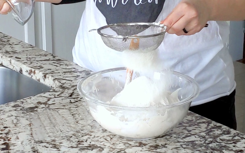 Hand holding a metal sifter shaking flour into a large glass bowl filled with a white mixture. Flour is falling from the sifter and is blurred in motion.