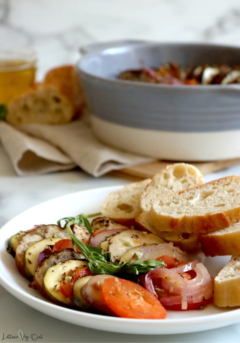 Plate with two rows of layered roasted vegetables topped with an herb sprig along with sliced baguette stacked on the plate to the right. Wood board with folded brown towel on it, topped with a grey and white round baking dish, baguette and small jar of olive oil sits blurred in the back.