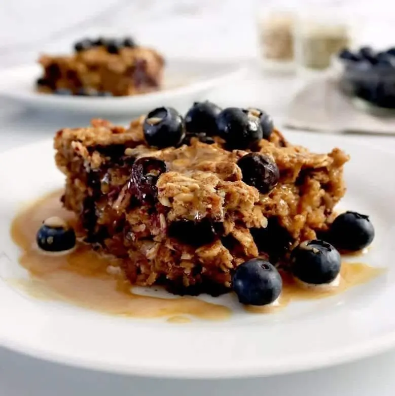 A served piece of vegan baked oatmeal served with blueberries and peanut butter drizzle. In the background is another piece of oatmeal and a small glass dish full of extra blueberries
