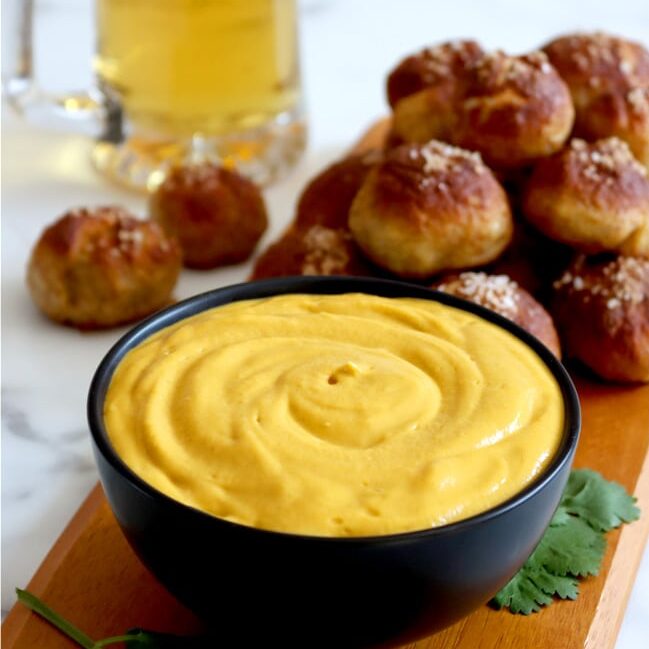 Black bowl filled with beer cheese dip sits on wood board with pile of pretzel bites stacked on the board behind the bowl. Glass mug of golden beer sits to the back left of the board with two pretzel bites in front of the mug. Green garnish around the bowl on the board.