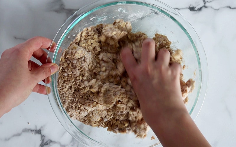 Hand crumbling butter into a mixture of oats, flour and brown sugar. Left hand holds the edge of the large glass bowl white the other hand is slightly blurred in motion of mixing.