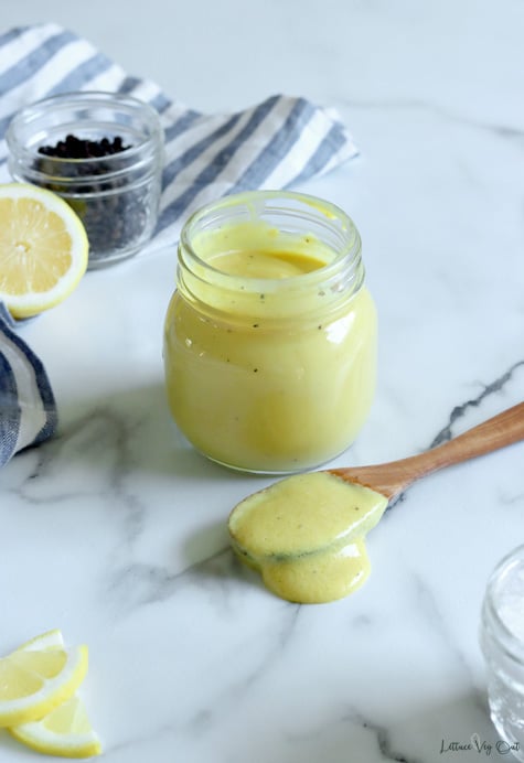 Small glass jar filled with creamy, light yellow sauce with wooden spoonful of the sauce placed in front of jar, on white-grey marble countertop. Lemon slices in bottom left corner with a blue and white stripped towel, half lemon and jar of peppercorns in top left corner.