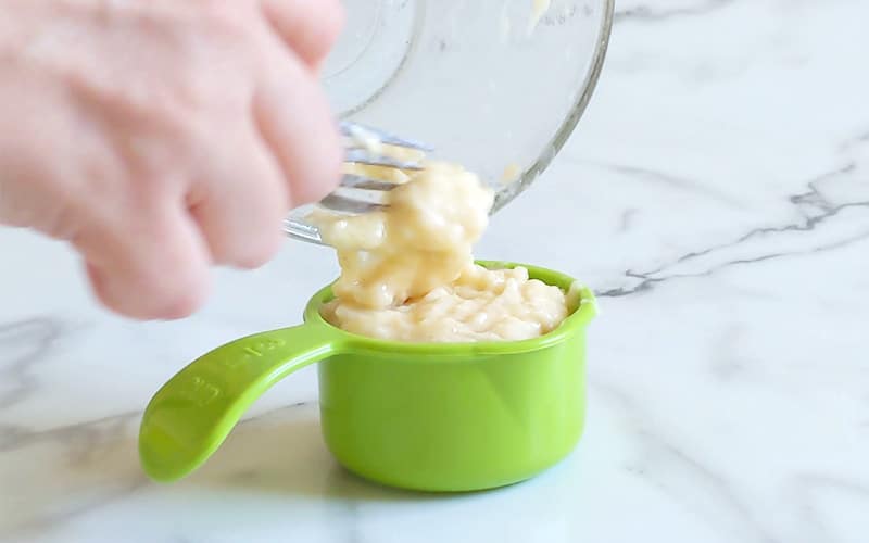Hand scooping mashed banana from glass bowl to green measuring cup using a fork. Hand is in top left corner and is slightly blurred, in motion.