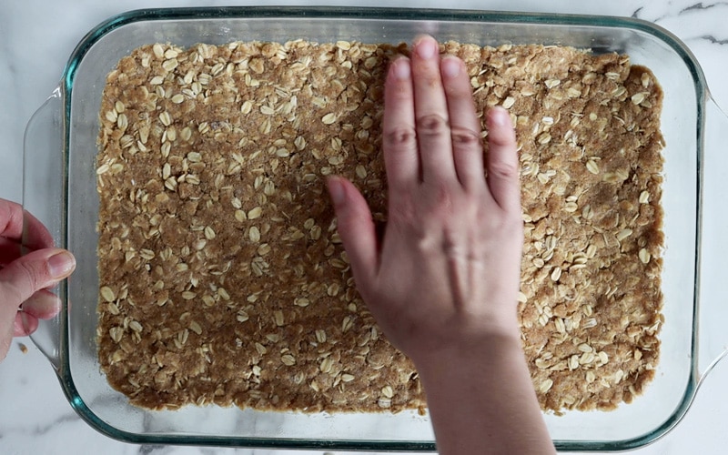 A hand presses a firm layer of oat crumble into the bottom of a large glass baking dish. The oat crumble appears firmly pressed and not crumby. A second hand is holding the baking dish on the left.