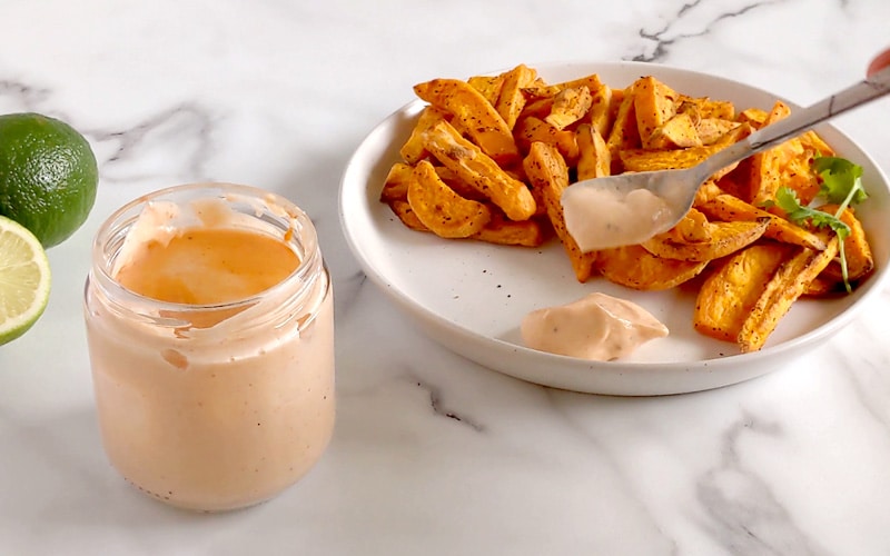 Spoon scooping sriracha mayo onto a round white plate full of sweet potato fries. A small glass jar of light orange mayo sits to the left of the plate with a whole and half lime further left.