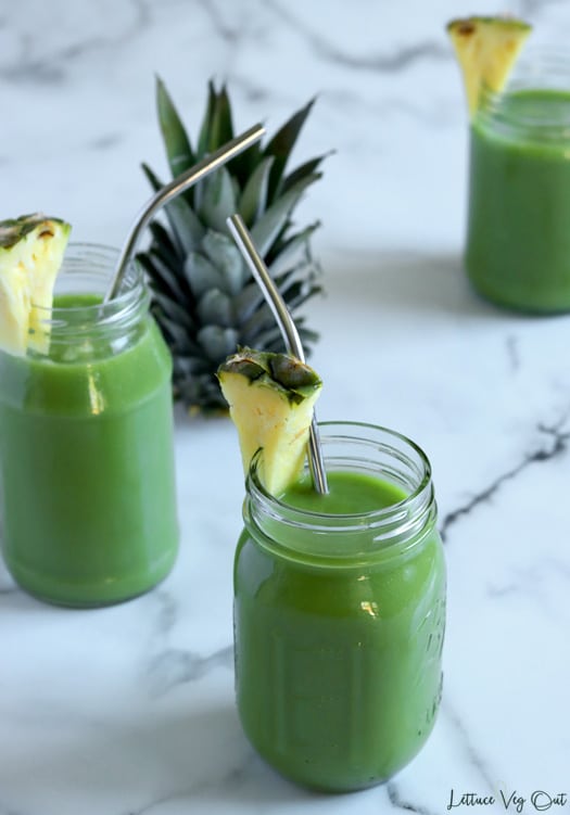 Arrangement of three glass jars filled with green juice and garnished with a piece of fresh pineapple sit on white-grey marble background. The top of a pineapple sits in the middle. The jars have bent metal straws placed in them.