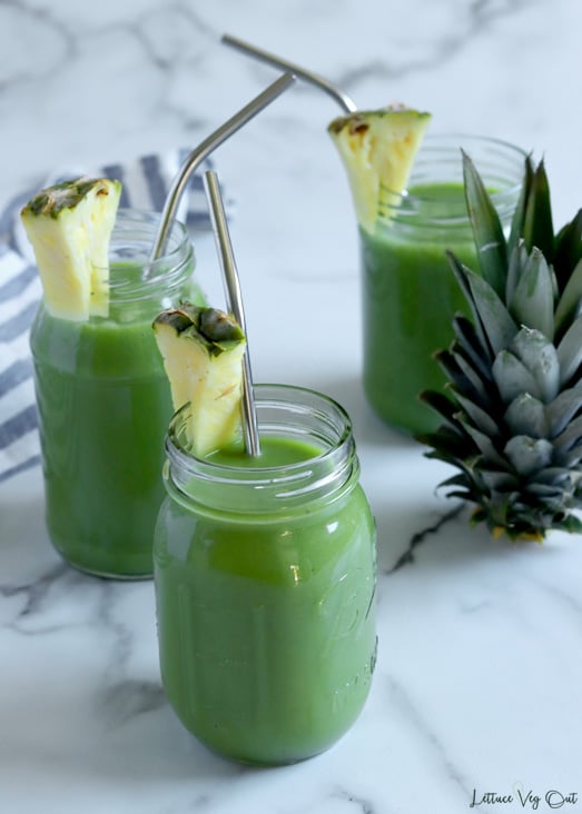 Three glass jars filled with green juice, garnished with pineapple and with metal straws sit on white-grey marble background with the top of a pineapple sitting to the right side of the image. White-blue stripped towel along the left edge.