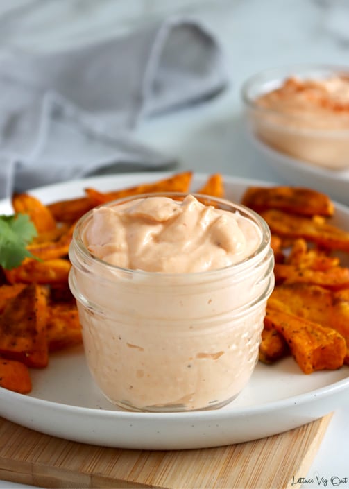 Small mason jar filled with spicy, light orange, mayonnaise on a white plate loaded with sweet potato fries. Plate sits on small light wood board and there's a grey towel blurred to the back left and second jar of mayo on a plate to the back right.