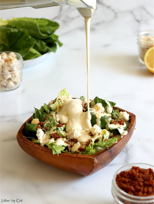 Square wood bowl filled with chopped lettuce, bacon bits and croutons with creamy Caesar dressing being poured from a blender onto the salad. Small jar of bacon bits in front right corner with a jar and lemon partly visible in the back right. Back left corner filled with plate of plain lettuce leaves and small jar of croutons.