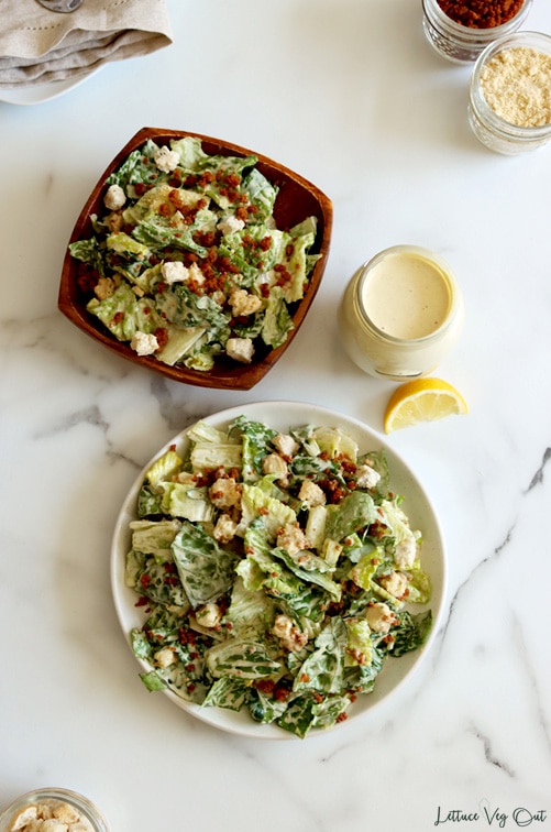 Top view of a white plate of tossed vegan Caesar salad (lettuce, bacon bits, croutons and creamy dressing) with a square wood bowl of salad above the plate which sit on a white-grey marble counter. Ingredients in the salad sit in jars around the plates and bowl.