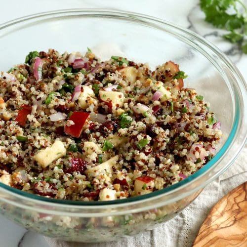 Square cropped image of large glass bowl filled with tri-color quinoa salad (white, red and black) made with chopped red apple, red onion, cilantro, walnuts and cilantro. Bowl sits on folded light brown towel over white-grey marble background with large wood spoon in front of bowl to the right and cilantro garnish in back right corner of image.