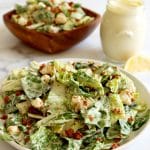 Close up of a plate of tossed Caesar salad (chopped Romaine lettuce, bacon bits and croutons with cream dressing). Blurred in the back is a jar of dressing with a lemon wedge and a square wood bowl filled with salad.