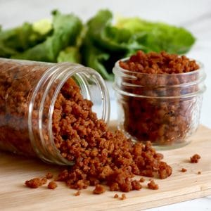Square cropped image of a Jar of bacon bits tipped on it's side with bacon crumbles falling out on a light wood board. Second small jar of bacon bits on the board to the back right. Lettuce blurred behind the jars, with a white-grey marble background.