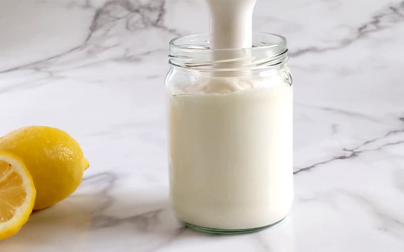Whipped mayonnaise (creamy white sauce) sits in a glass jar with an immersion blender handle placed inside the jar (handle visible coming out of jar). White-grey marble background with two lemons to the left of the jar.