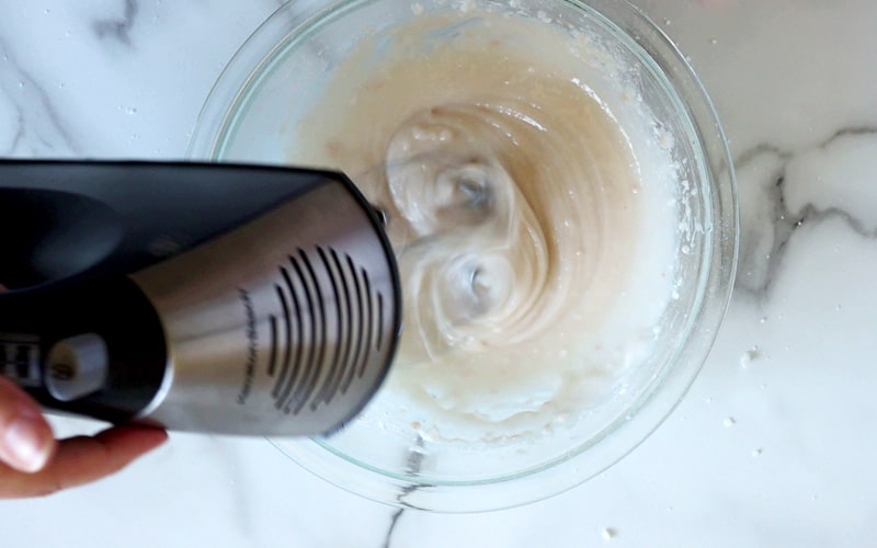 A hand holds a metal and black electric beater that is whipping off-white (very light brown) frosting in a glass bowl. Frosting appears glossy and has a few flecks of vanilla and icing sugar throughout.