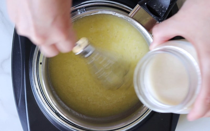Top view of a pot filled with melted butter. One hand holds a small whisk and is whisking the sauce in the pot while another hand holds a small glass jar of milk, about to pour it into the pot.