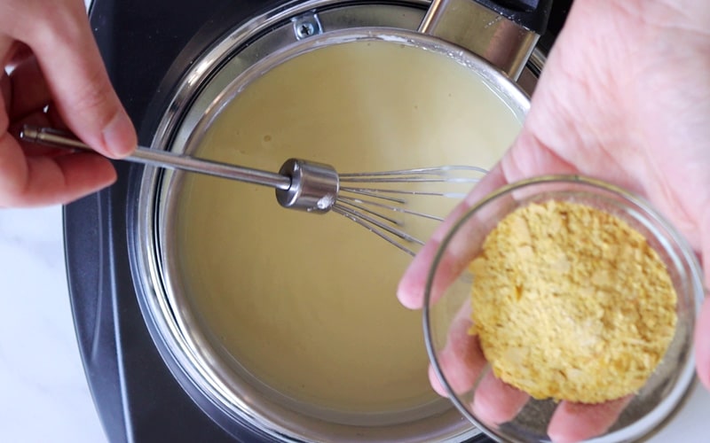 Top view of a pot with a light yellow, creamy sauce in it. A hand on the left side of the image holds the end of a small whisk that is sitting in the sauce. A second hand, on the right side of the image, holds a small glass dish full of nutritional yeast, over the pot.