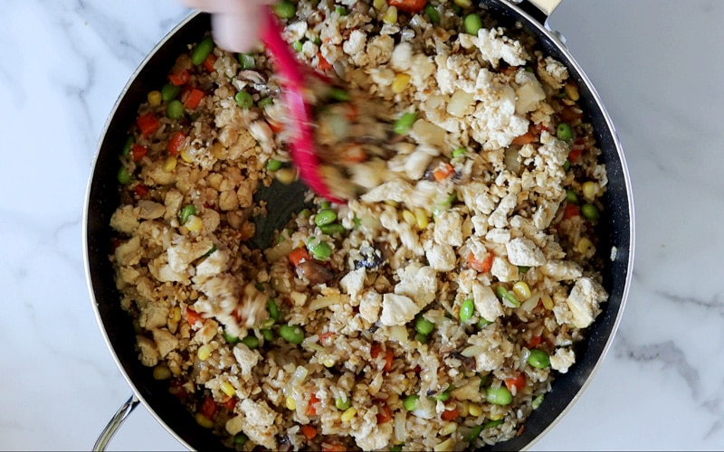 Top view of a large pan filled with tofu fried rice (crumbled tofu, rice, corn, carrots, edamame, mushrooms) with a large red spoon tossing the food around in the top-center of image. A bit of a hand is visible holding the spoon which is blurred in motion.