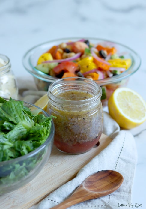 Glass jar filled with Greek salad dressing sitting on light wood board with a glass bowl of chopped lettuce to the left of the jar. A second glass bowl filled with colorful chopped vegetables sits off the wood board behind the jar of dressing, sitting on a white-grey marble background