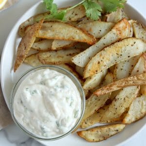 Square cropped image of the top view of a round white plate topped with golden brown potato wedges and a small glass jar filled with garlic mayo that is creamy and white with flecks of green onion. Plate is garnished with a sprig of cilantro.