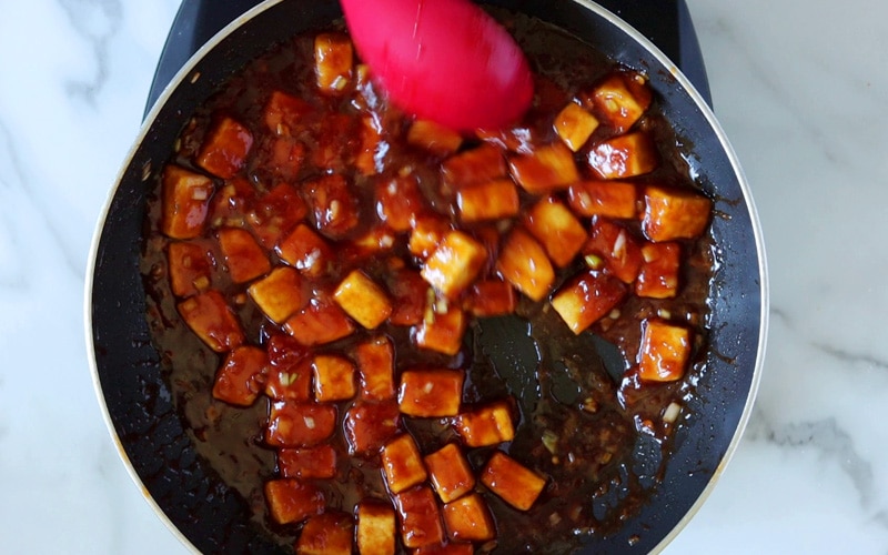 Top view of a pan of tofu cubes coated in a thick General Tso sauce. Some of the bottom of the pan is visible and a large red spoon hovers over the pan in the top center of image.