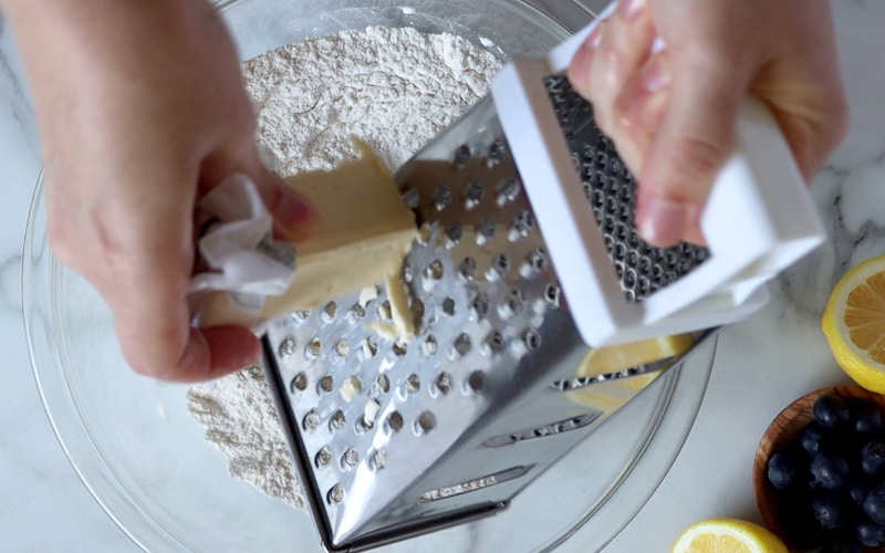 A hand hold the handle of a metal box grater over a glass mixing bowl full of flour. A second hand holds a stick of butter and is grating the butter. Blueberries and lemon sit in the bottom right of image.