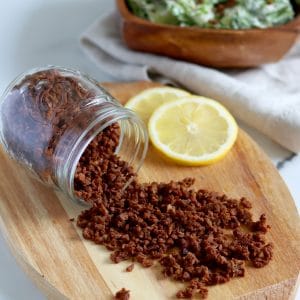 Square cropped image of a jar of veggie bacon bits sits on its edge on an oval wood board with lots of bacon bits spilling out of jar, all over the wood board. Two slices of lemon are on the board to the back right.