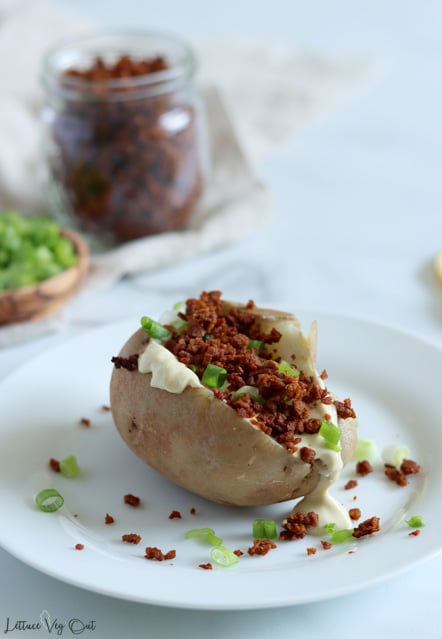 Baked potato on round white plate, with a cut down the center of the potato and topped with creamy, white colored sauce (Caesar dressing), chopped green onion and bacon bits. The bacon bits and green onion are spilled over the potato all over the plate. Blurred jar of bacon bits and small wood bowl of chopped green onion blurred in back left of image.