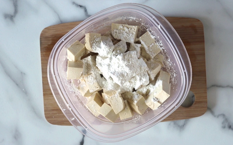 Top view of a plastic container filled with tofu cubes and a pile of corn starch on top of the tofu. Container sits on a small wood board with a white-grey marble background.