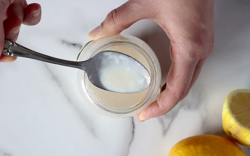 Top view of a small jar filled with milk that a hand is holding in place while a second hand holds a spoonful of thick and slightly chunky over the top of the jar.