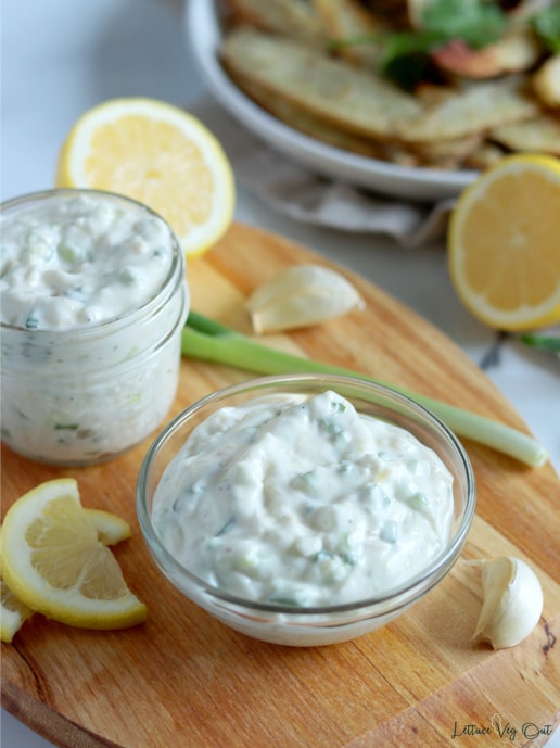 Close up of a small glass bowl filled with creamy garlic mayo that is white with flecks of green onion. Bowl sits on a wood board with a second small glass jar of mayo behind it. Board is decorated with lemon slices, a green onion and some garlic cloves. A plate of potato wedges and a half lemon sit in the back right corner of image.