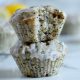 Square cropped image of a stack of two lemon poppy seed muffins with drizzled glaze over them with the top muffin having a large bite taken from it. Blurred muffins and lemons in the background.