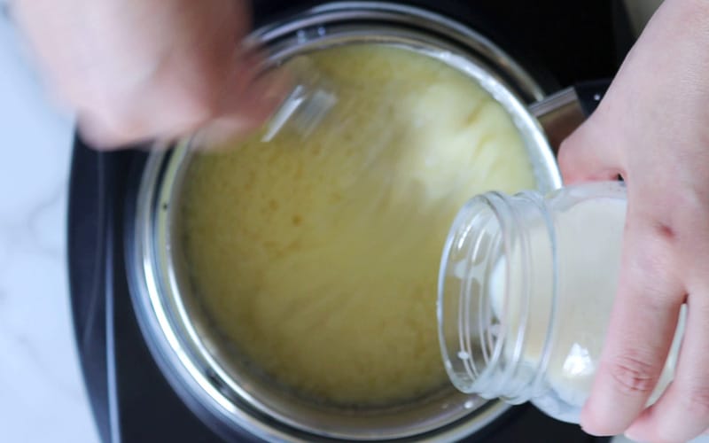 Top view of a pot that contains a light yellow mixture that is being whisked by a hand holding a whisk at the top edge of the pot. The contents of the pot are blurred, in motion. A second hand is pouring milk into the pot from the right side of the image. The sauce looks like there are some areas of melted butter and some smooth and creamy sections.
