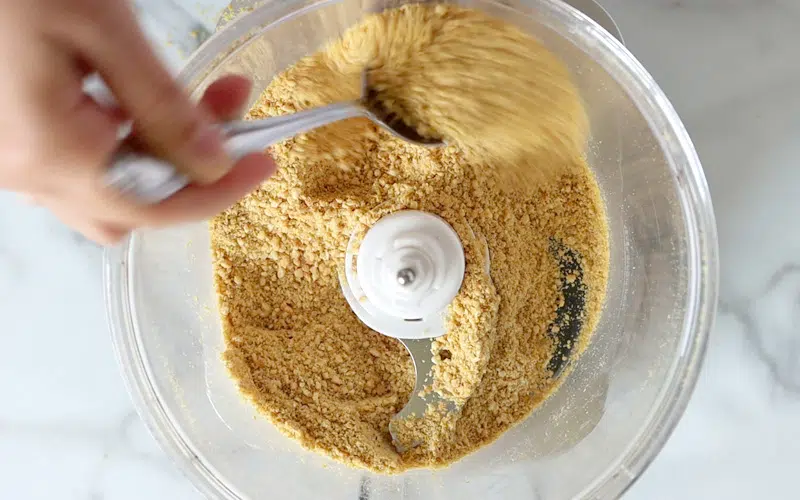 Top view of a food processor filled with a crumbly cashew mixture. A hand holding a metal spoon stirs the mixture in the top portion of the image; hand and spoon are blurred in motion.