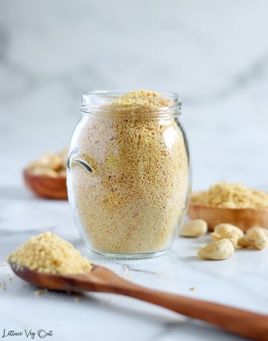 Straight on view of a small glass jar filled with vegan cashew parmesan cheese with a wood spoonful of the crumbly parmesan sitting in front of the jar. Some cashews sit to the right of the jar with a small wood dish of additional cashews blurred in the back right corner and a second wood dish blurred further back.