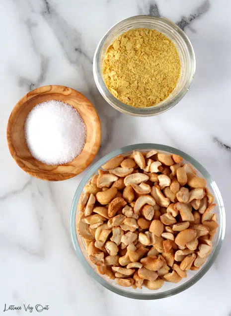 Top view of a small glass jar filled with nutritional yeast, a small wood dish filled with salt and a larger glass bowl filled with cashew pieces, all sitting on white-grey marble.