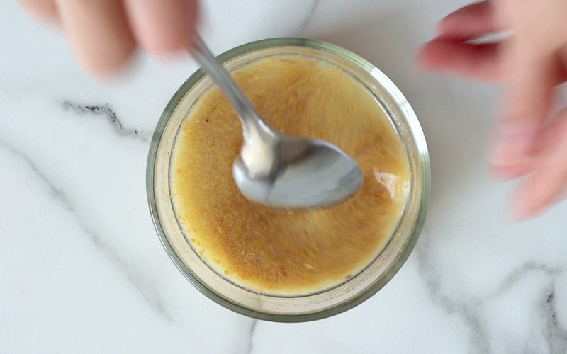 Top view of a small glass dish filled with ground flaxseed that is being mixed with lemon juice. A hand holding a spoon is blurred, stirring the flaxseed.