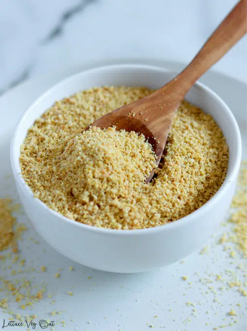 Small white bowl filled with crumbly vegan parmesan cheese (ground cashews) with a wood spoon resting in the bowl. Bowl sits on plate with the parmesan dusted on the right and left side.