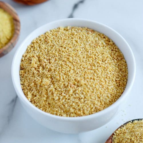 Square cropped image of a small white bowl filled with vegan cashew parmesan (golden brown-yellow crumbly mixture) sitting on white-grey marble.