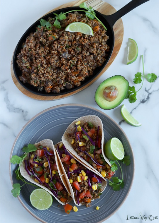 Top view of an oval cast iron pan sitting on a wood board and filled with quinoa and lentil taco "meat" at the top of image. Bottom half of image shows a grey-blue plate topped with three prepared tacos filled with quinoa taco "meat", tomato, corn, red cabbage and cilantro with cilantro and lime garnishes on the plate and between the plate and cast iron pan on the marble background.