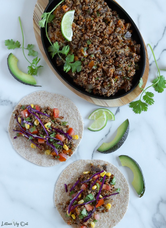 Top view of two tacos sitting flat on a marble background and topped with quinoa and lentil mixture, tomato, corn, red cabbage and cilantro with a cast iron pan on an oval wood board filling the top half of the image. Cast iron filled with cooked quinoa taco filling. Avocado slices, lime wedges and cilantro sprigs places around the tacos and pan.