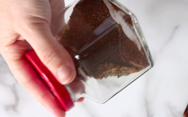 Hand holding a square glass jar with bright red lid that has dark red and brown spices in it. Hand and jar are slightly blurred, in motion as the jar is being shaken.
