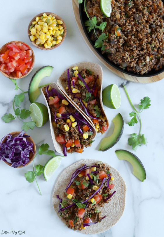 Top view of two folded quinoa tacos in the center, one flat quinoa taco at the bottom of image and a cast iron pan filled with quinoa taco filling partially cropped out from the top right corner. Small wood dishes filled with red cabbage, tomato and corn places along left edge of image along with avocado slices, lime wedges and cilantro surrounding the tacos for garnish.
